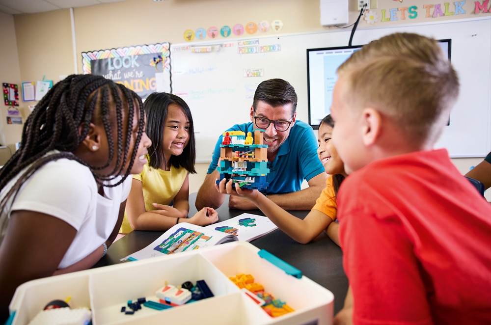 Ein Lehrer und mehrere Schüler bewundern gemeinsam ein großes LEGO-Modell im Klassenzimmer.