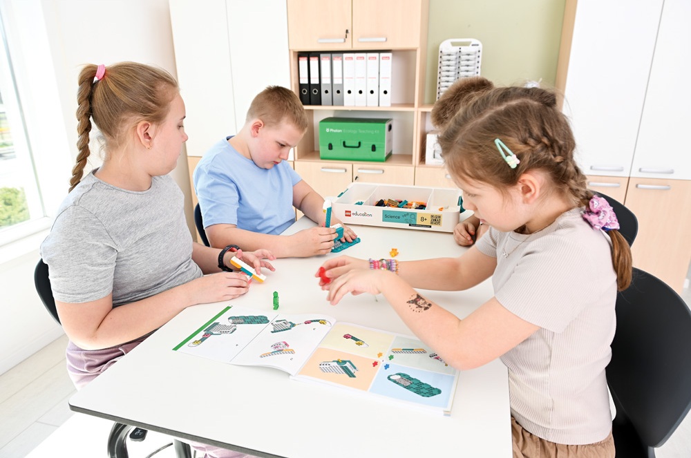Vier Kinder bauen LEGO-Modelle anhand von Anleitungen an einem Tisch im Klassenzimmer.